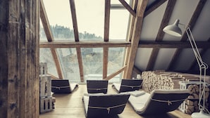Acogedor dormitorio en ático con vigas de madera, ventanas amplias y vistas a árboles. Hay sillones de mimbre, una lámpara y una pila de leña.