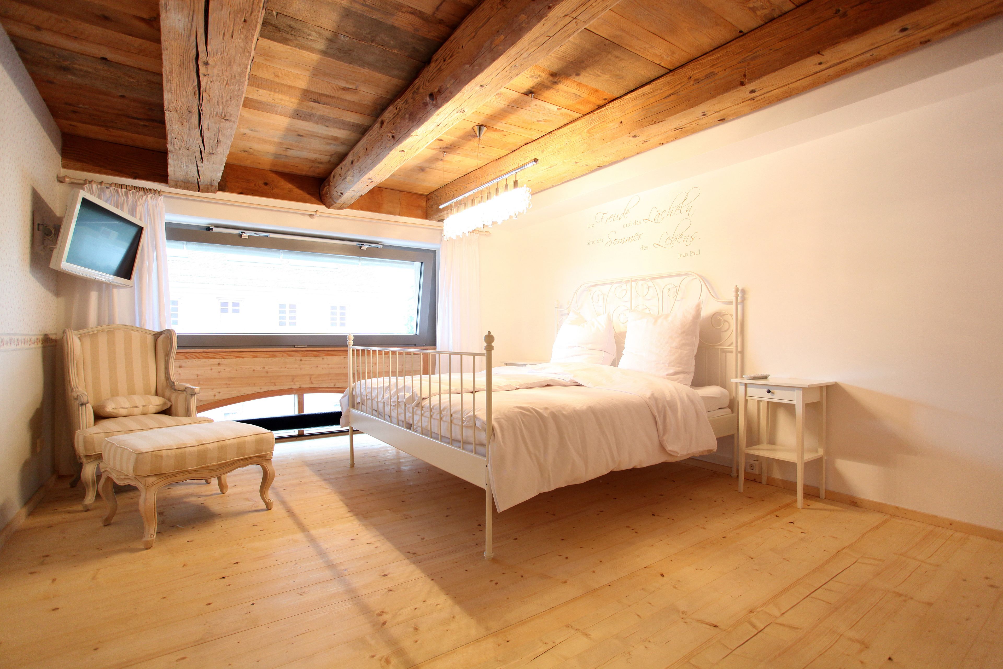 Ein Schlafzimmer mit Holzbalkendecke, einem Bett, einem Stuhl, einem Fernseher und einem Fenster.