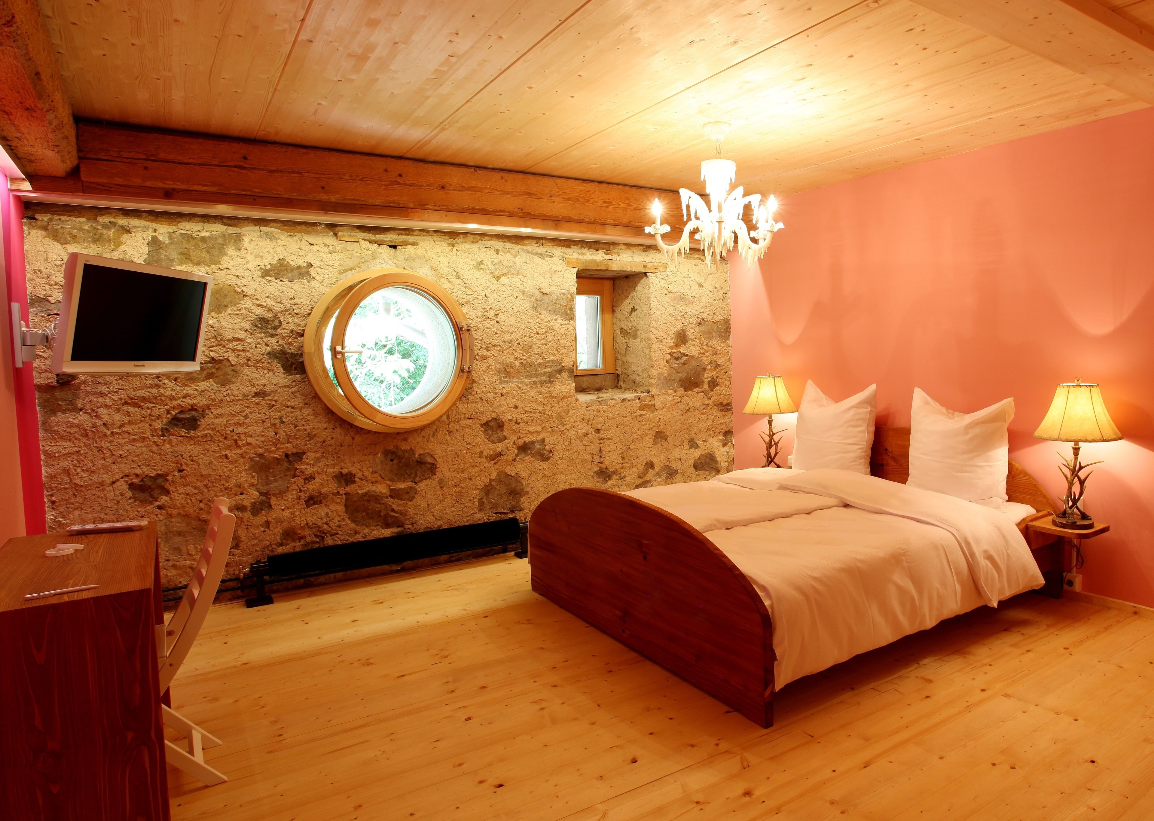 A bedroom with a wooden bed, a round window, a stone wall, and a television mounted on the wall.