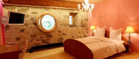 A bedroom with a wooden bed, a round window, a stone wall, and a television mounted on the wall.