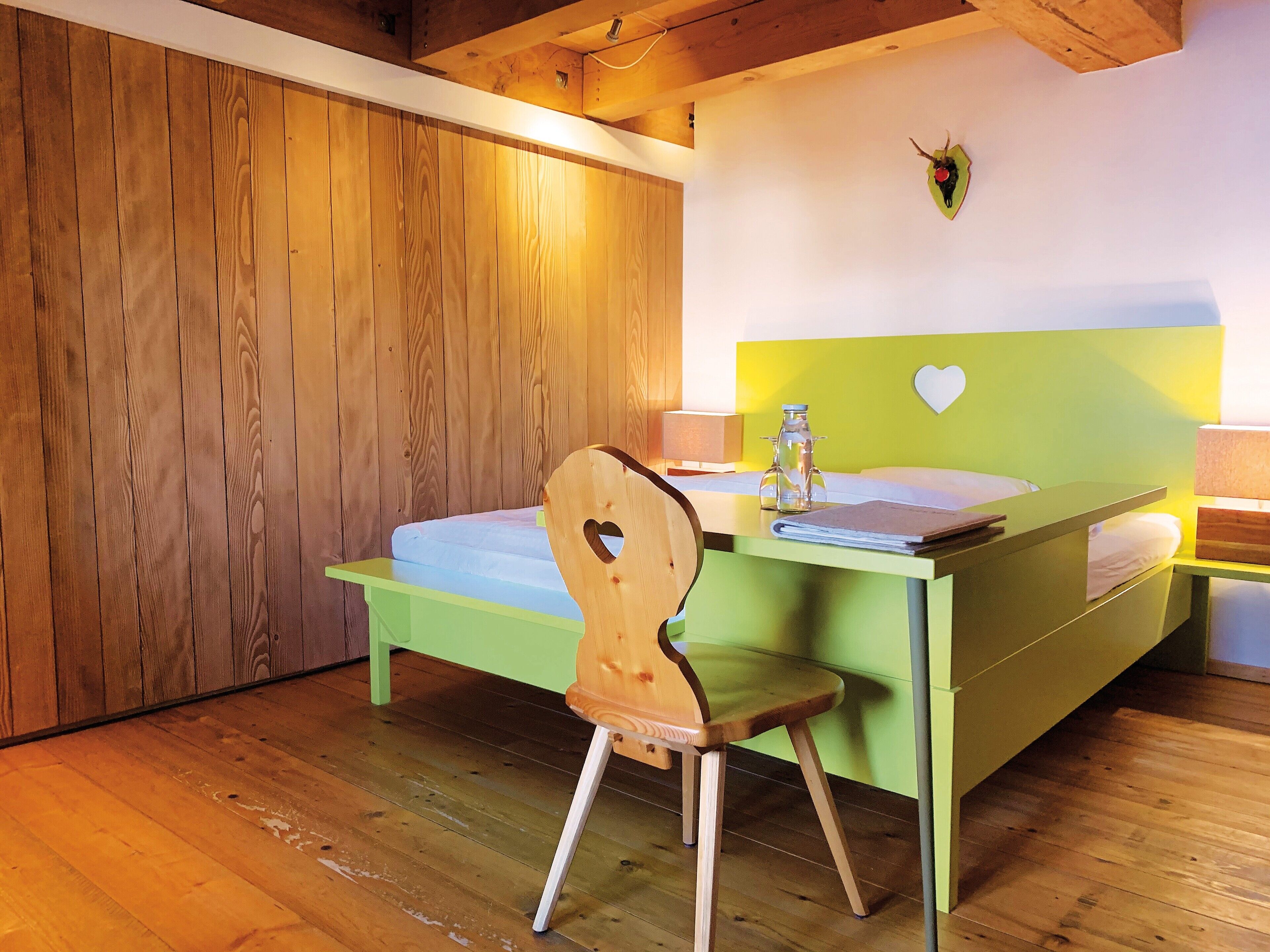 Una habitación con suelo de madera, una cama con cabecera verde, una silla de madera con un corazón recortado y una lámpara fijada en la pared.