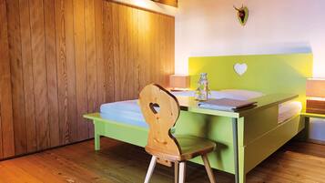 Una habitación con suelo de madera, una cama con cabecera verde, una silla de madera con un corazón recortado y una lámpara fijada en la pared.