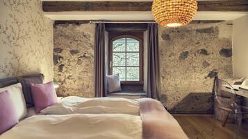 Un dormitorio con pared de piedra, una ventana con cortinas, una cama con ropa blanca y una silla transparente.