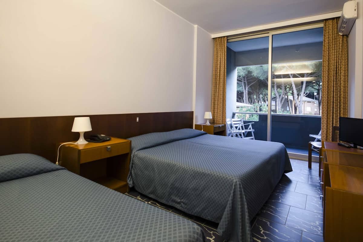 Une chambre d’hôtel avec deux lits, un bureau, une chaise, une télévision et un balcon donnant sur des arbres et des immeubles.