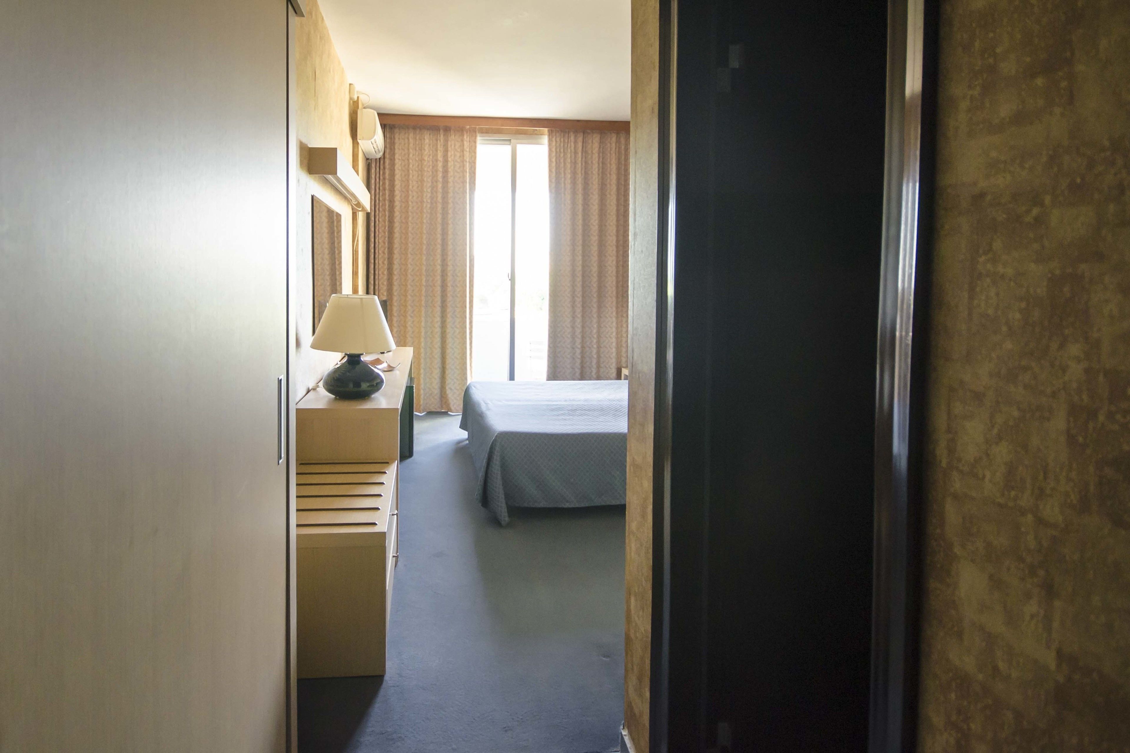 Une chambre d’hôtel avec un lit, un bureau doté d’une lampe, une commode et une fenêtre avec des rideaux.