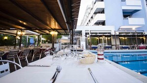 An outdoor dining area with tables set for a meal, a pool, and a modern building in the background. - Hotel Helios (Tarquinia)