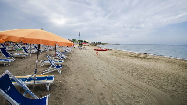 A beach with rows of sun loungers and umbrellas, a lifeguard tower, and a red boat on the sand. - Hotel Helios (Tarquinia)
