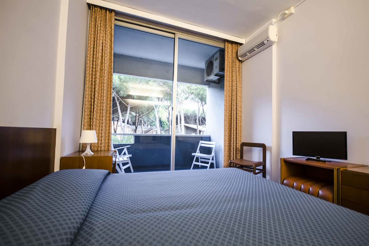 Une chambre d’hôtel avec un grand lit, un bureau avec une télévision, une chaise et un balcon donnant sur des arbres et un immeuble.