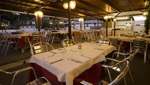 An outdoor dining area with tables set for multiple guests, featuring metal chairs and a covered roof structure. - Hotel Helios (Tarquinia)