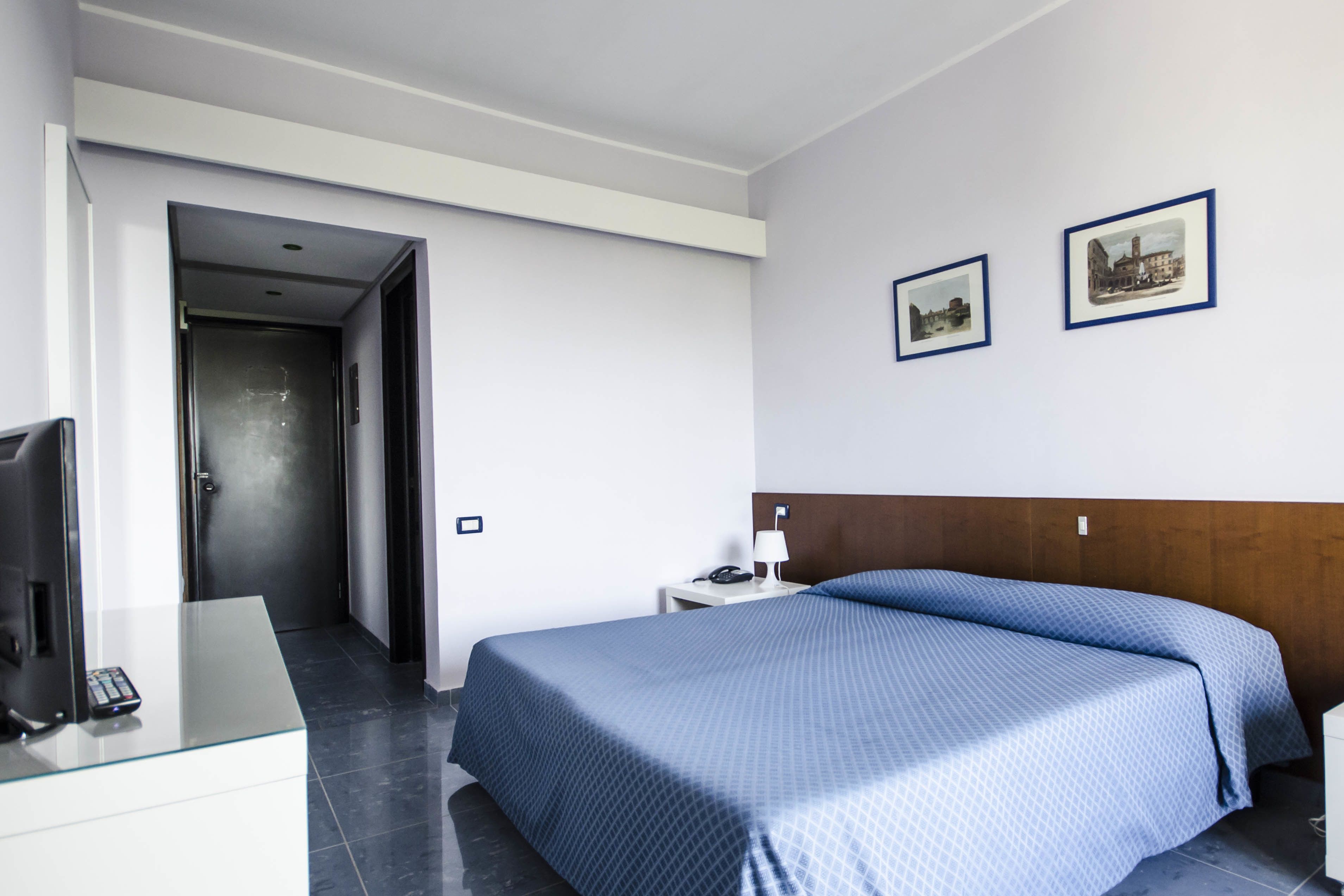 Habitación de hotel con cama, escritorio con computadora, lámpara y dos cuadros en la pared.