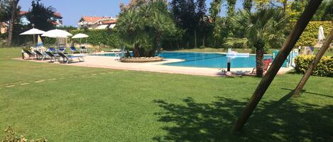 A pool area with a grassy lawn, lounge chairs, and umbrellas.