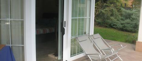 A room with a bed, a table with a blue tablecloth, and a sliding glass door leading to a patio with two lounge chairs.