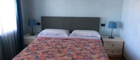 Una cama bien hecha con una colcha estampada con motivos florales, dos lámparas de noche, una mesita de noche y un cuadro de un paisaje sobre la cama.