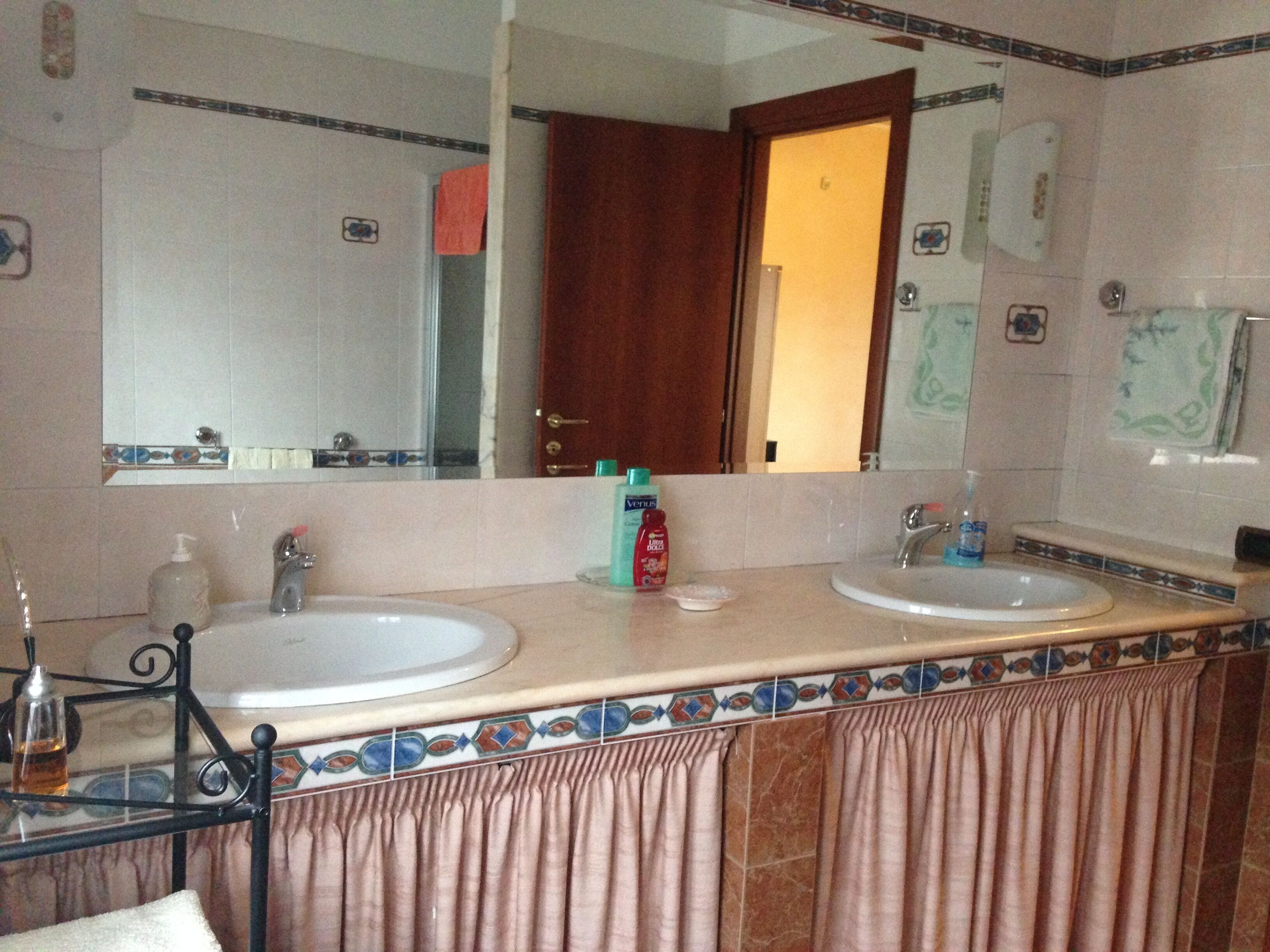 Un baño con dos lavabos, un espejo y una puerta que da a otra habitación.