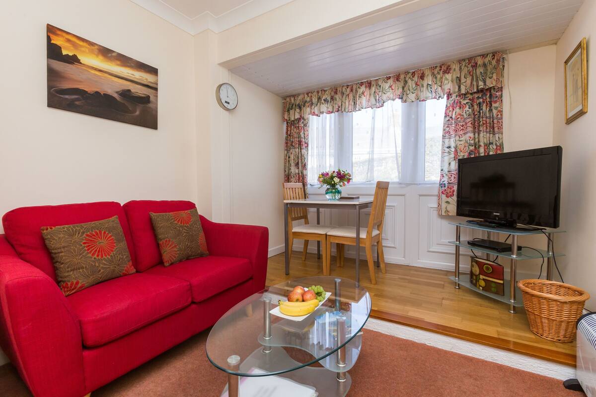 A living room with a red sofa, a glass coffee table with fruit, a TV on a stand, and a window with floral curtains.