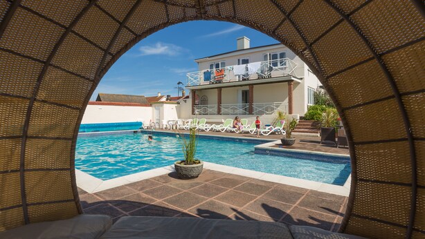 A pool area with a clear view through an archway, a pool, lounge chairs, and a building with a balcony in the background. - Beau Vallon Holiday Apartments (St. Pierre du Bois)