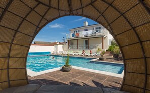 A pool area with a clear view through an archway, a pool, lounge chairs, and a building with a balcony in the background.