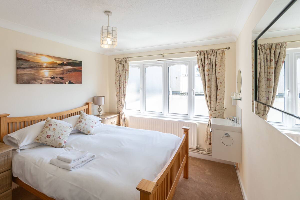 A bedroom with a bed, a nightstand, a lamp, a window with curtains, a radiator, and a painting on the wall.