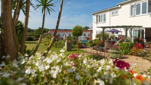A garden with palm trees and flowers, a pool, and a building with a covered patio. - Beau Vallon Holiday Apartments (St. Pierre du Bois)