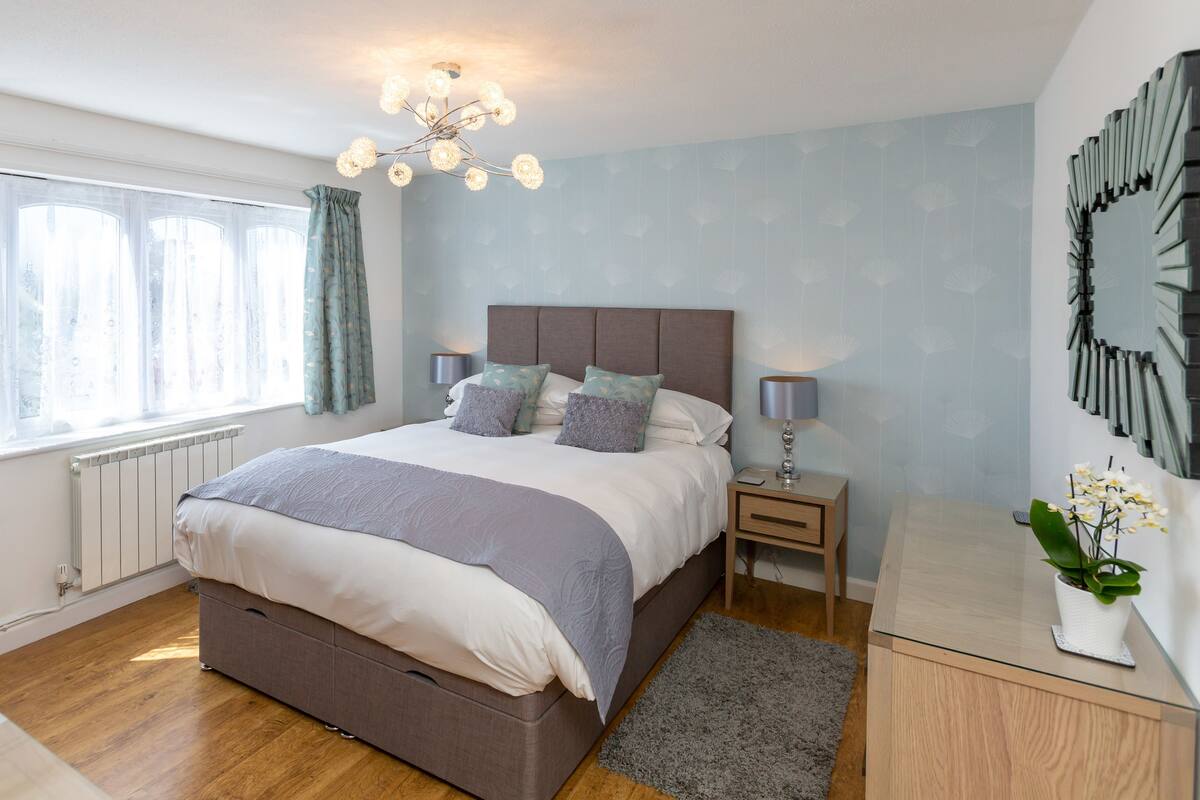 A bedroom with a large bed, bedside tables, a radiator, a window with curtains, a chandelier, and a wall-mounted mirror.