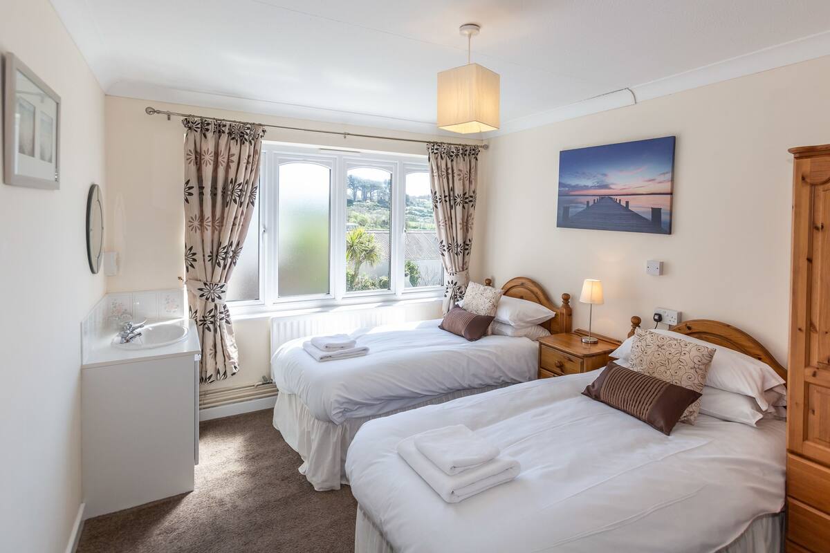 A bedroom with two beds, a wooden wardrobe, a bedside table with a lamp, a window with curtains, and a painting on the wall.