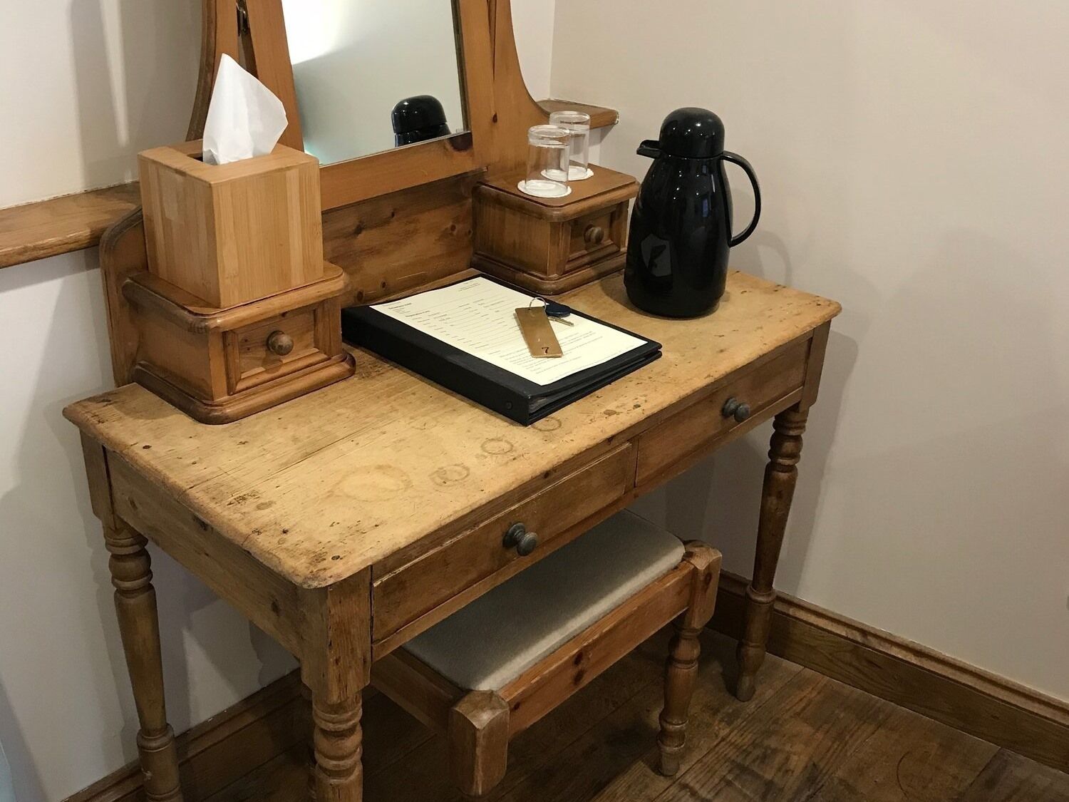 Un bureau en bois avec un miroir, une boîte de mouchoirs, une cafetière noire et un présentoir à documents.