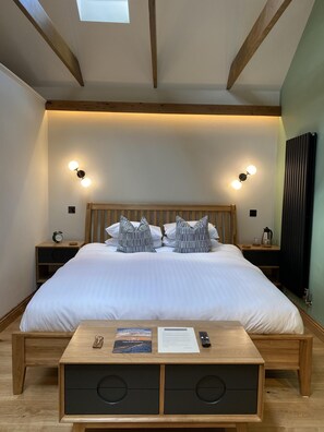 A neatly arranged bedroom with a large bed, two bedside tables, a wooden nightstand, a bedside lamp, a small table with a book and a remote control, and a skylight above the bed. - Sebastians (Oswestry)