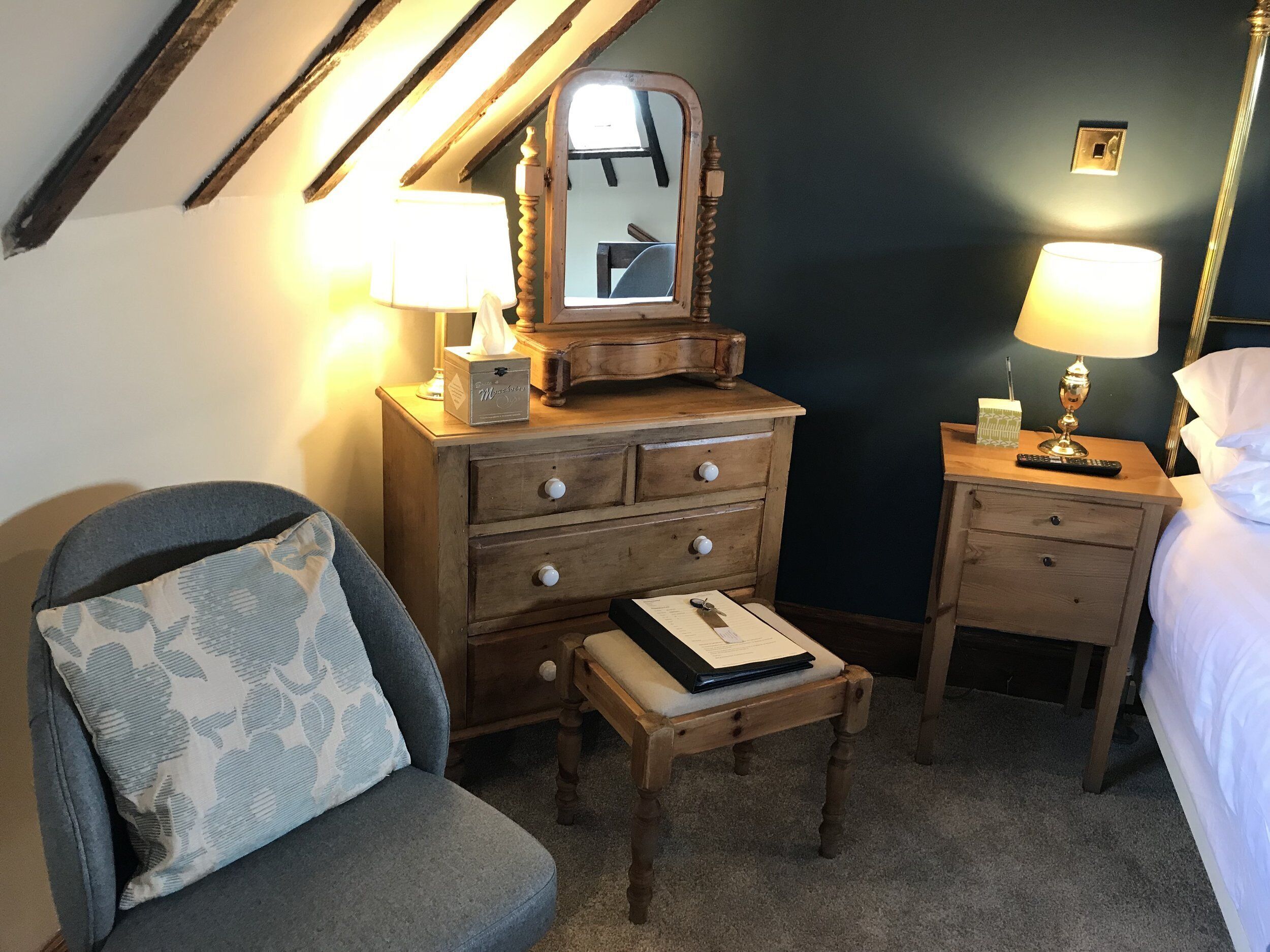 A bedroom with a wooden dresser, a bedside table with a lamp, a chair with a cushion, and a small table with a folder.