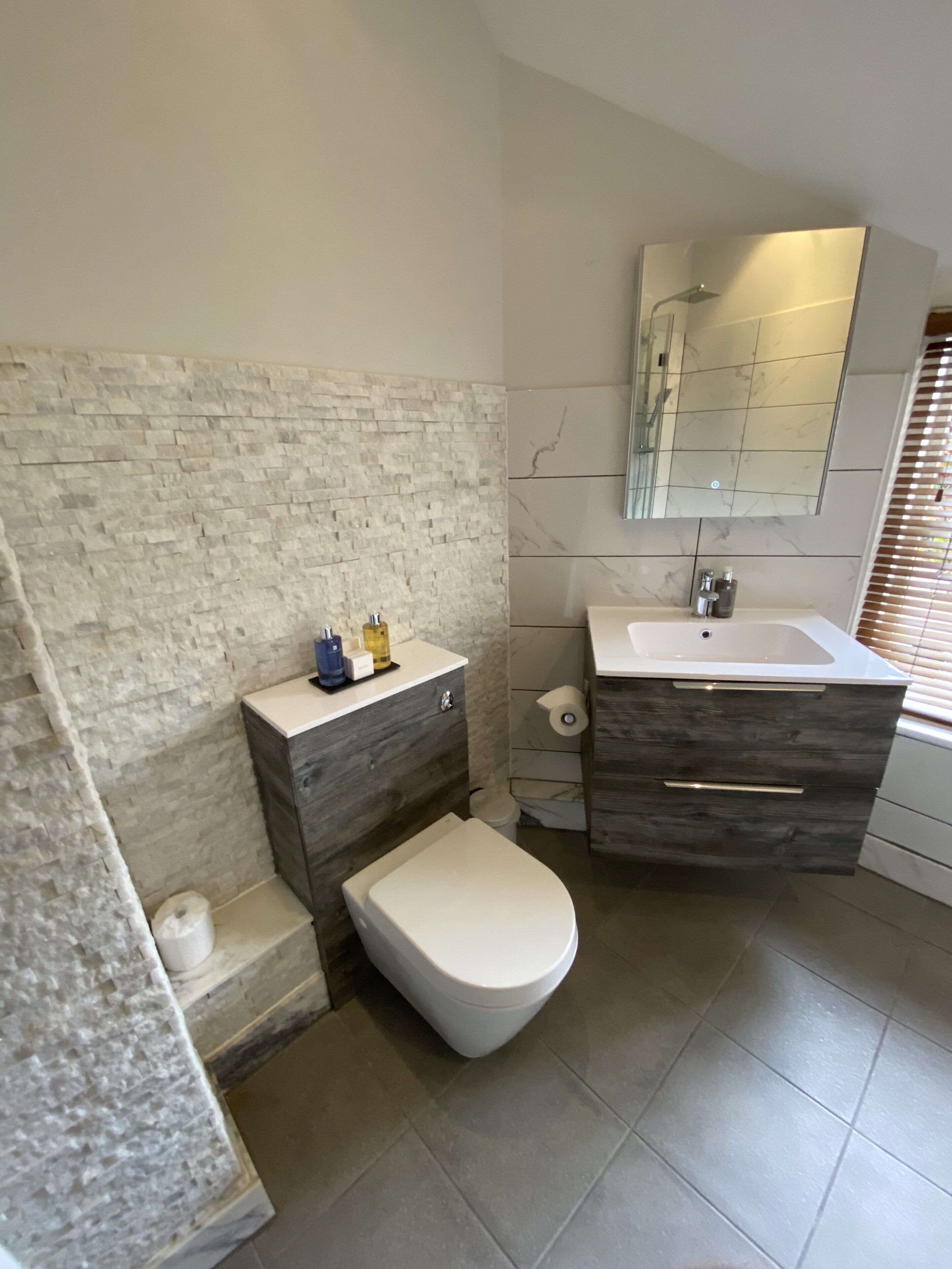 Un baño moderno con pared de piedra, inodoro blanco, tocador con espejo y zona de ducha.