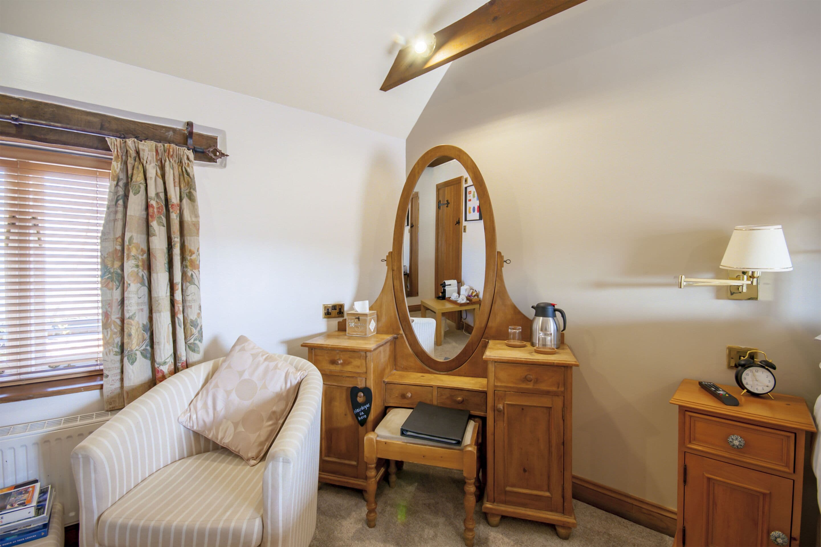 Un dormitorio con cómoda de madera, espejo, mesita de noche con lámpara, silla y ventana con cortinas.