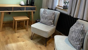 Una habitación moderna con un escritorio de madera, dos sillones grises, un espejo redondo, un televisor de pantalla plana y una ventana con persianas.