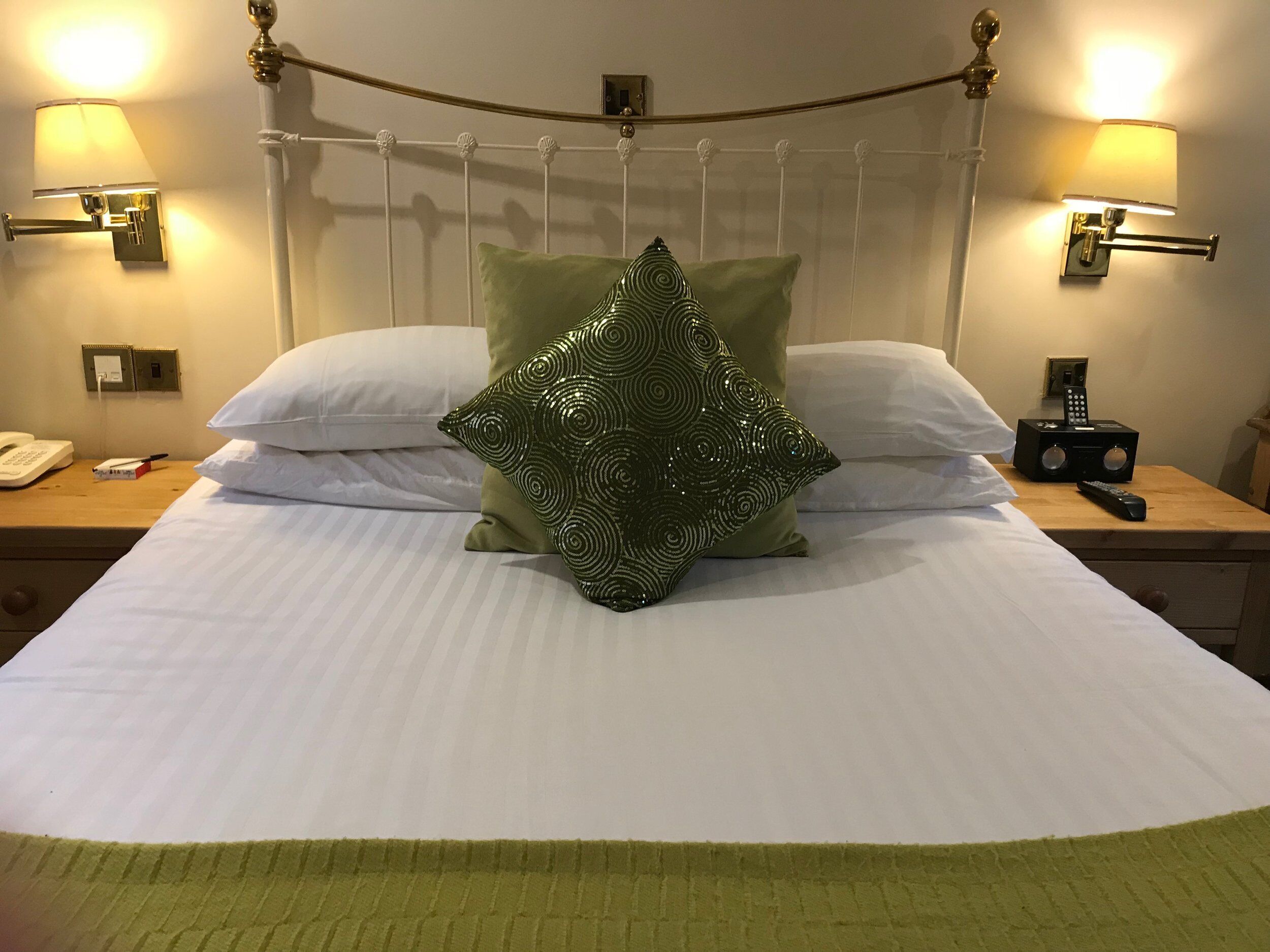 A neatly made bed with a green patterned pillow, white pillows, and a green throw at the foot of the bed. There are two bedside tables with lamps and a telephone on one.