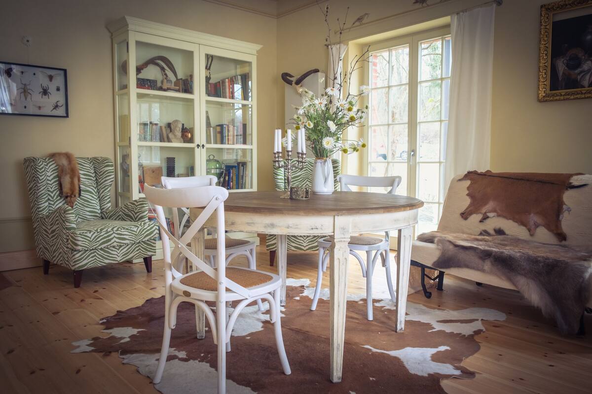 A cozy living room with a round wooden table, white chairs, a zebra-patterned armchair, a cowhide rug, a bookshelf, and a window with curtains.