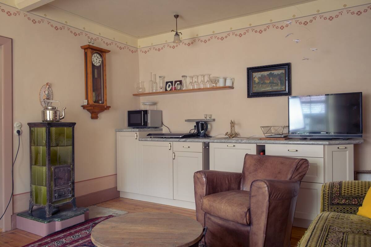 A compact living space with a kitchenette, a brown leather chair, a patterned sofa, a wooden clock, and a framed painting on the wall.
