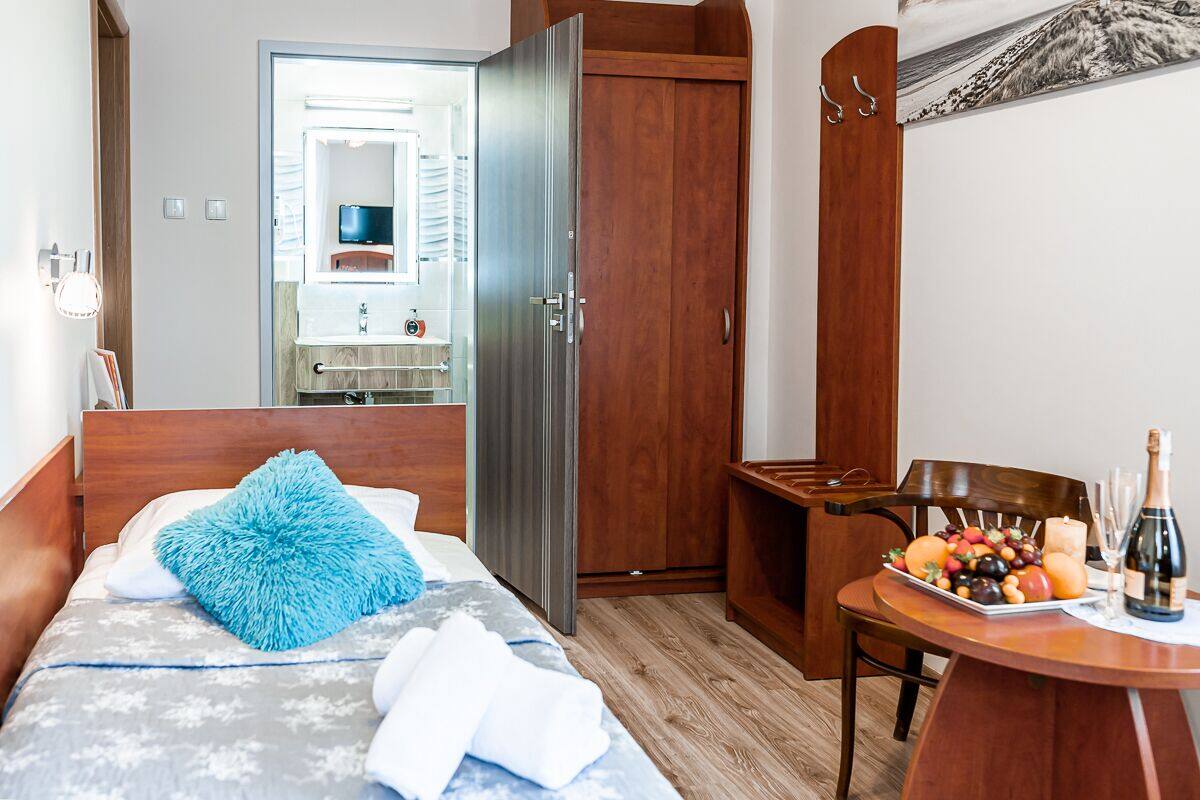 Una habitación de hotel con una cama, un armario de madera, una mesita con una bandeja de frutas y una botella, y un espejo al fondo.