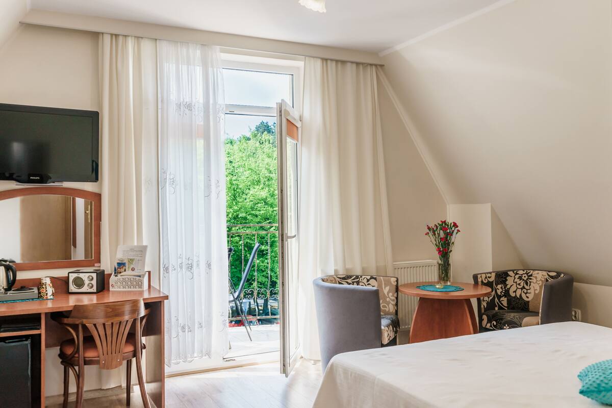 Habitación de hotel con cama, escritorio con radio y teléfono, una silla, mesita con un jarrón de flores y balcón con vistas al verdor.