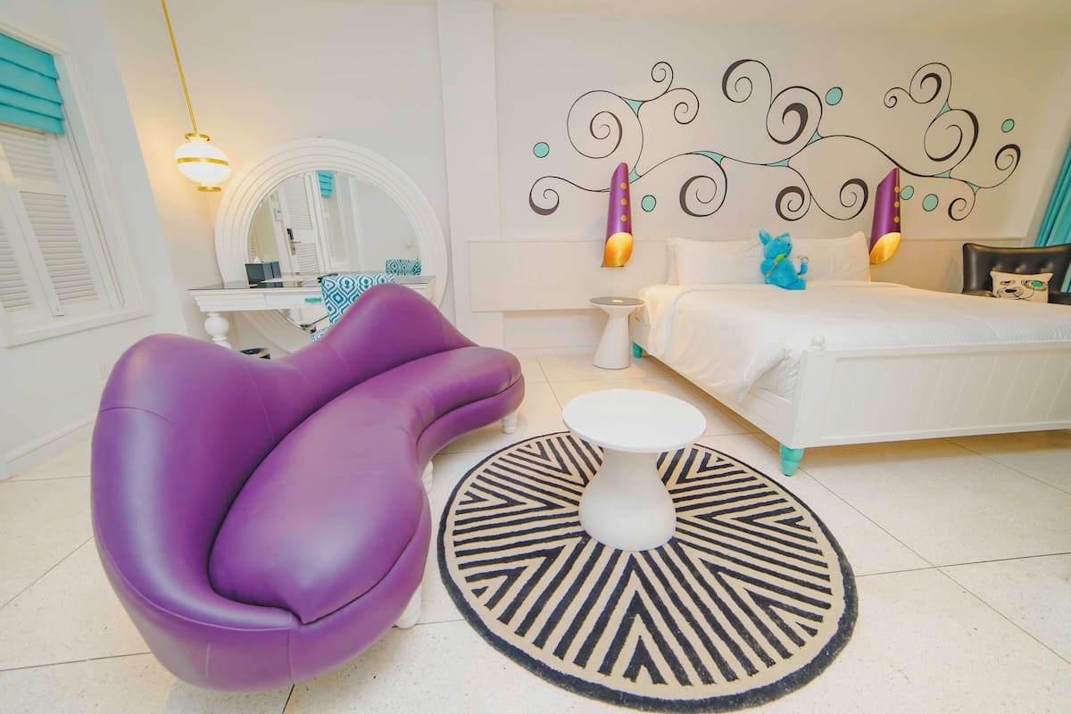 A modern bedroom with a purple armchair, a round coffee table, a bed with white bedding, a decorative wall, and a mirror.
