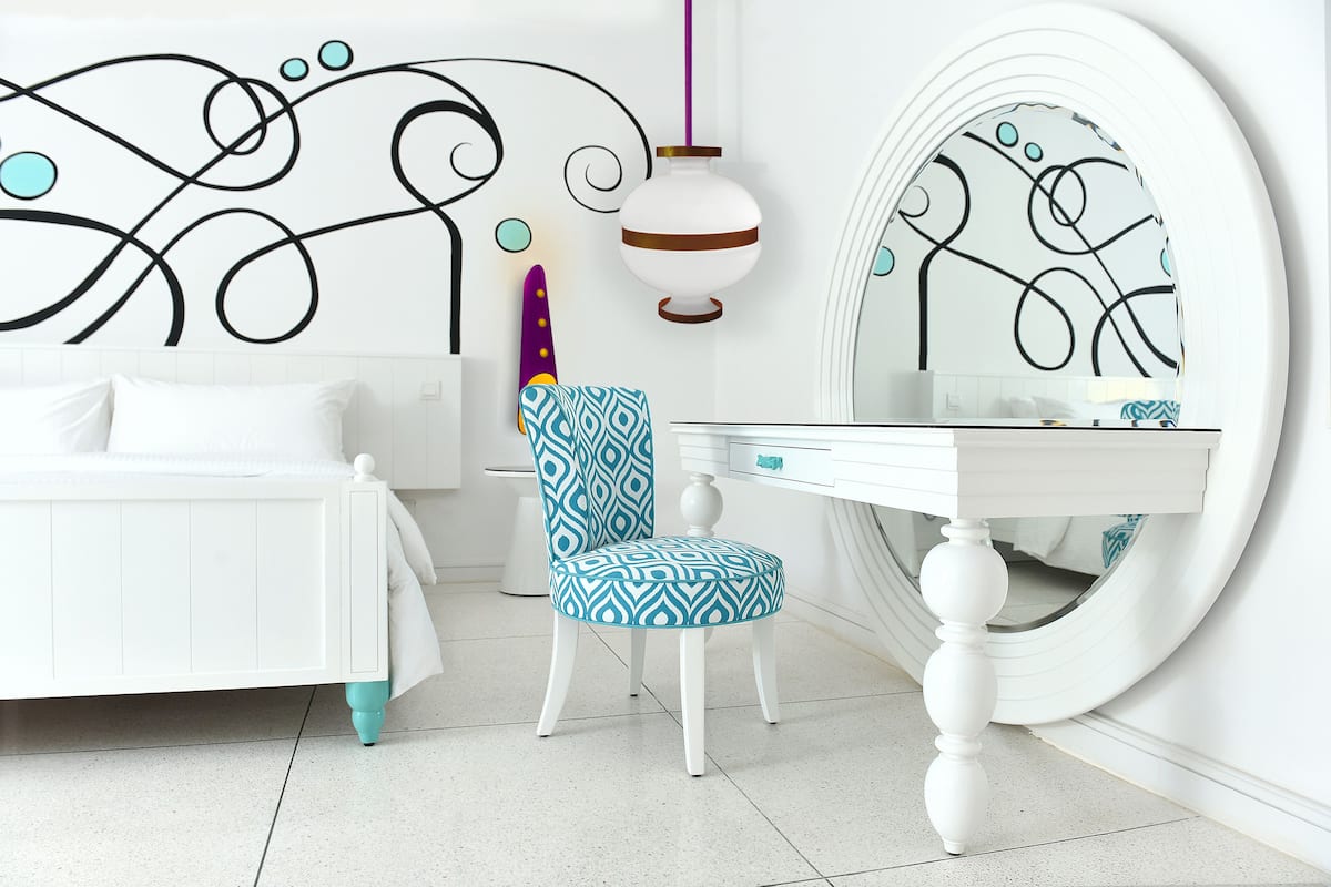 A bedroom with a bed, a chair, a desk, a mirror, and a wall with a decorative design.