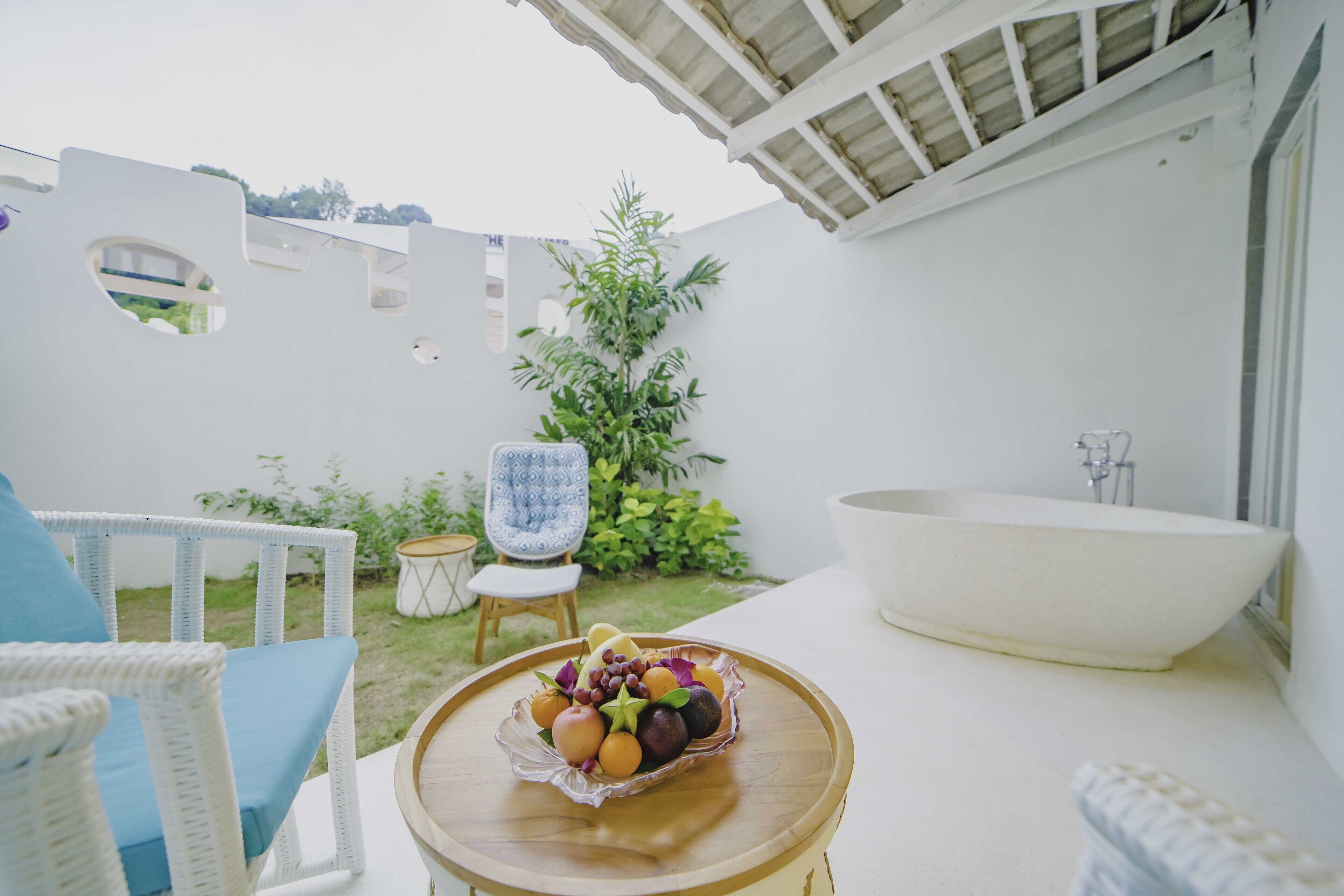 A bathroom with a freestanding bathtub, a small round table with a fruit bowl, and a wicker chair with a cushion.