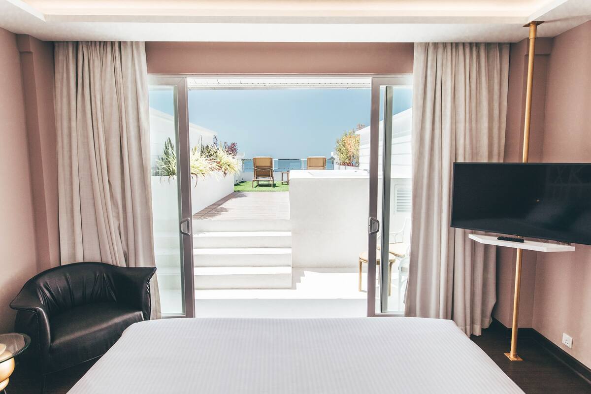 A hotel room with a bed, a TV, a black leather chair, and a balcony with a view of the sea.