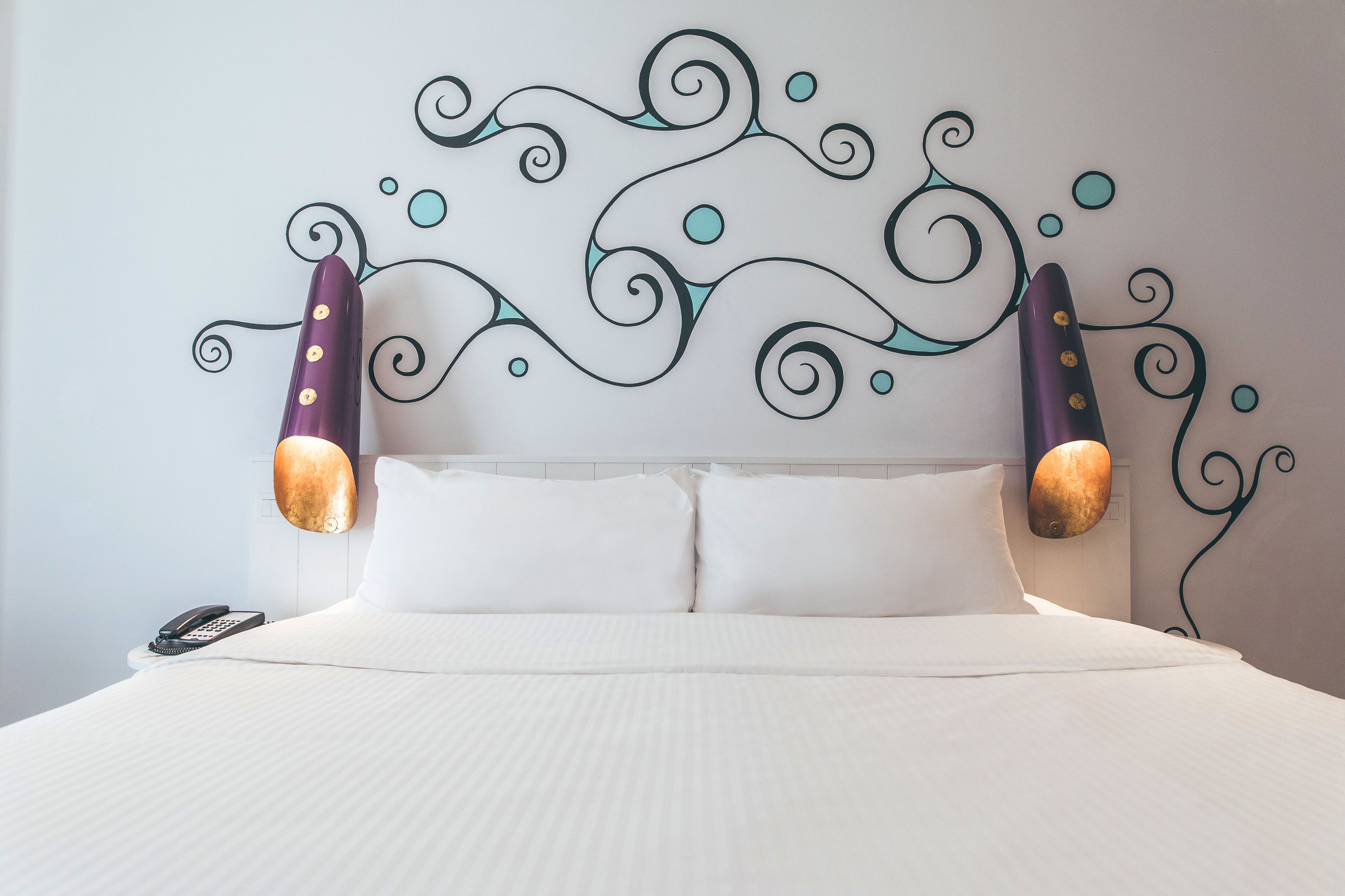 A neatly made bed with white linens, a wall-mounted telephone, and decorative wall art with swirling patterns and circular motifs.