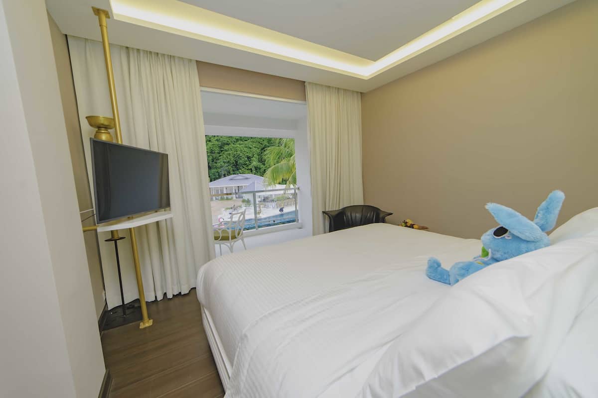 A hotel room with a large bed, a television on a stand, a view of a beach and palm trees through the window, and a plush toy on the bed.