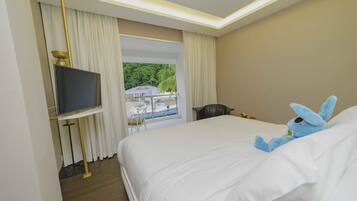A hotel room with a large bed, a television on a stand, a view of a beach and palm trees through the window, and a plush toy on the bed.