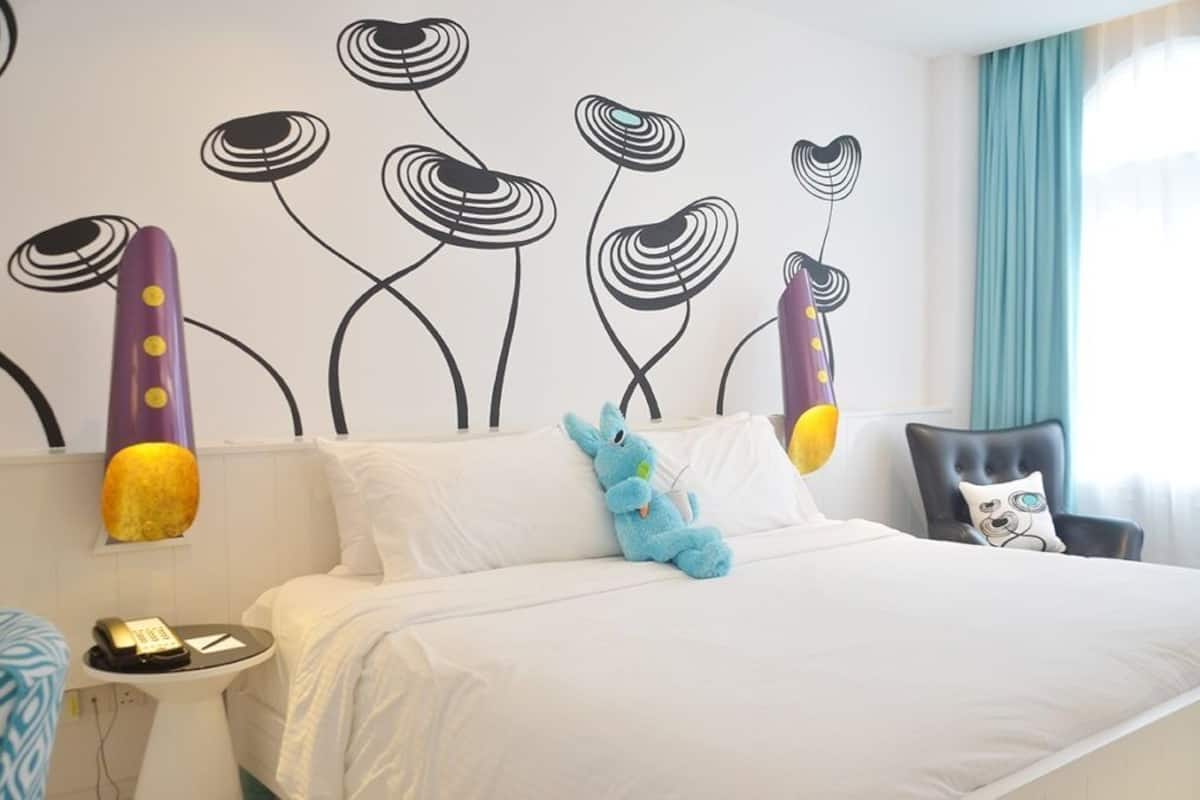 A hotel room with a large bed, a wall mural, a blue plush toy, a chair, and a bedside table with a phone.