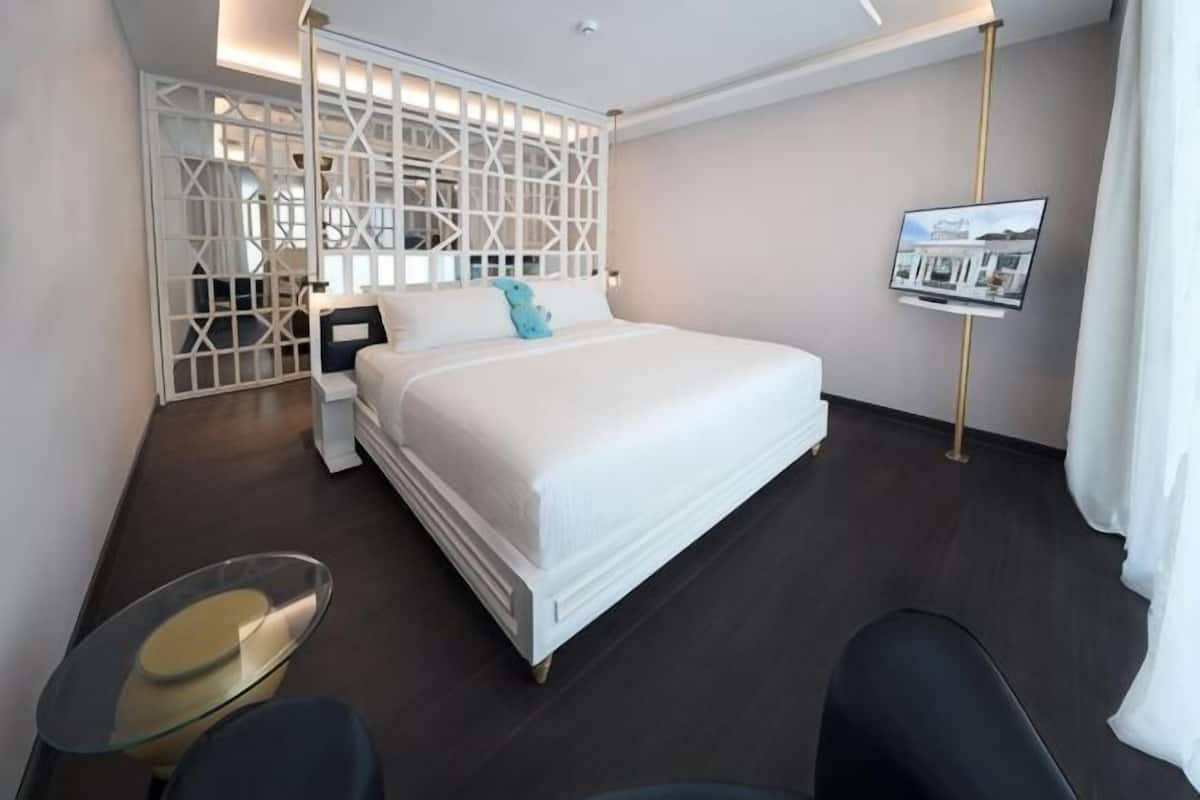 A hotel room with a bed, a glass table, a chair, a TV, and a decorative partition.