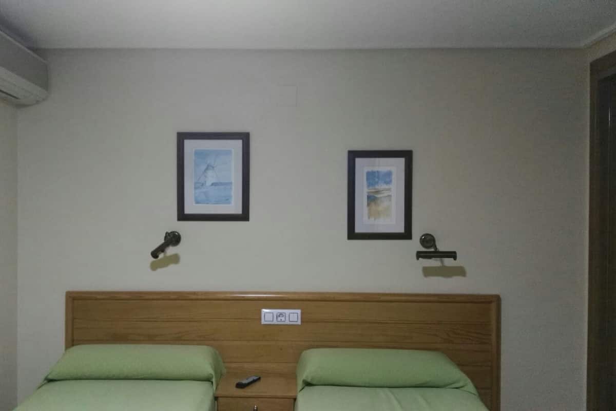 A room with two single beds, a wooden headboard, two framed pictures on the wall, and a wall-mounted air conditioner.