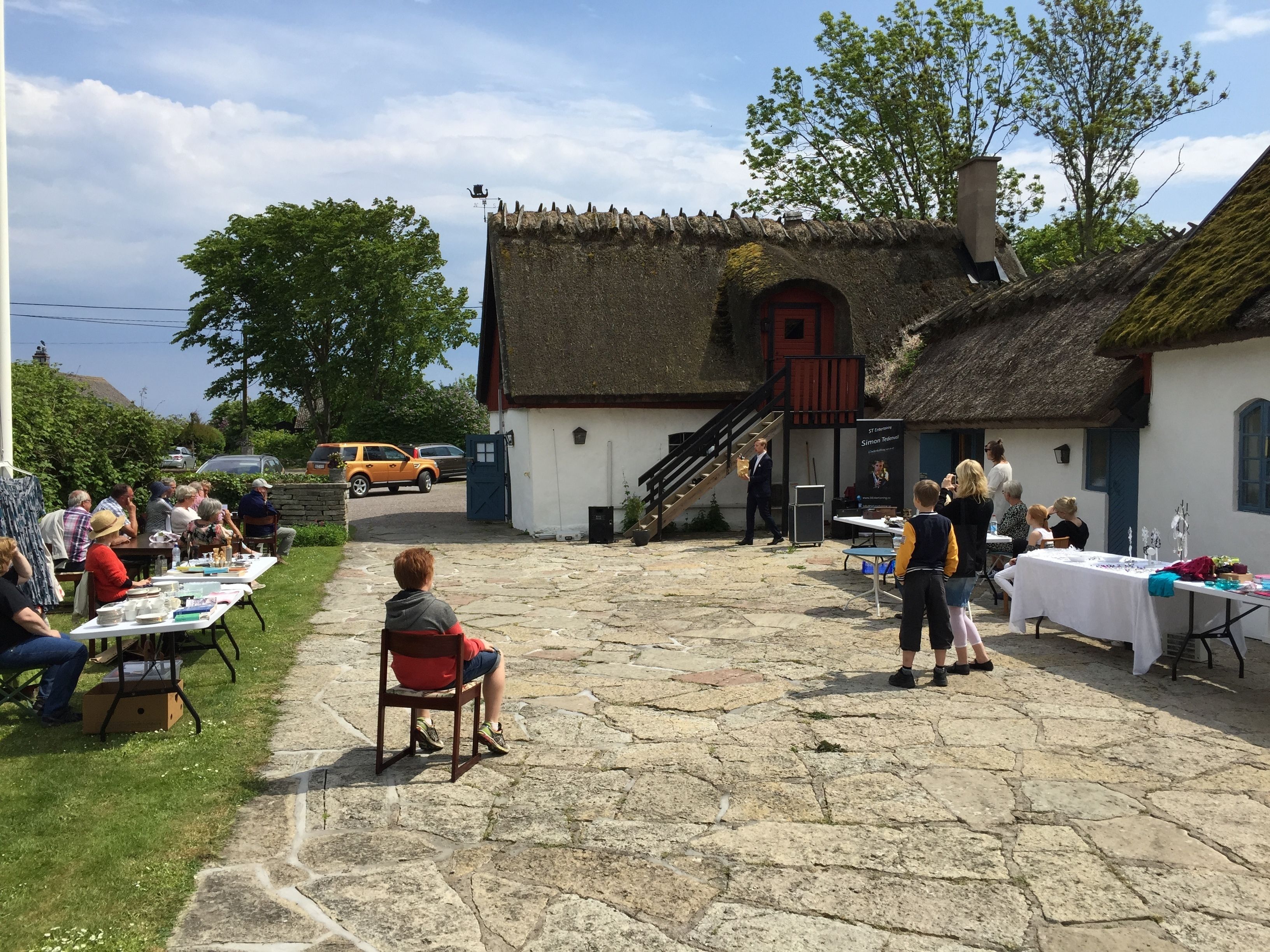 An outdoor event with people seated at tables, a building with a thatched roof, and a staircase leading to a door.