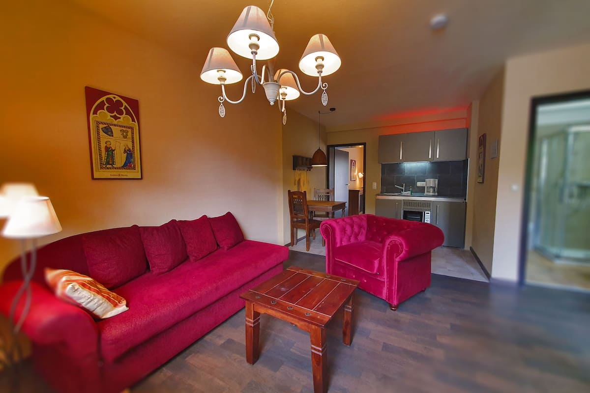 Een woonkamer met een rode bank, een houten salontafel en een kroonluchter. In de achtergrond is een keuken met moderne apparatuur te zien.