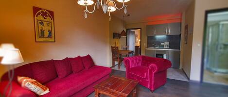 A living room with a red sofa, a wooden coffee table, and a chandelier. There is a kitchen in the background with modern appliances.