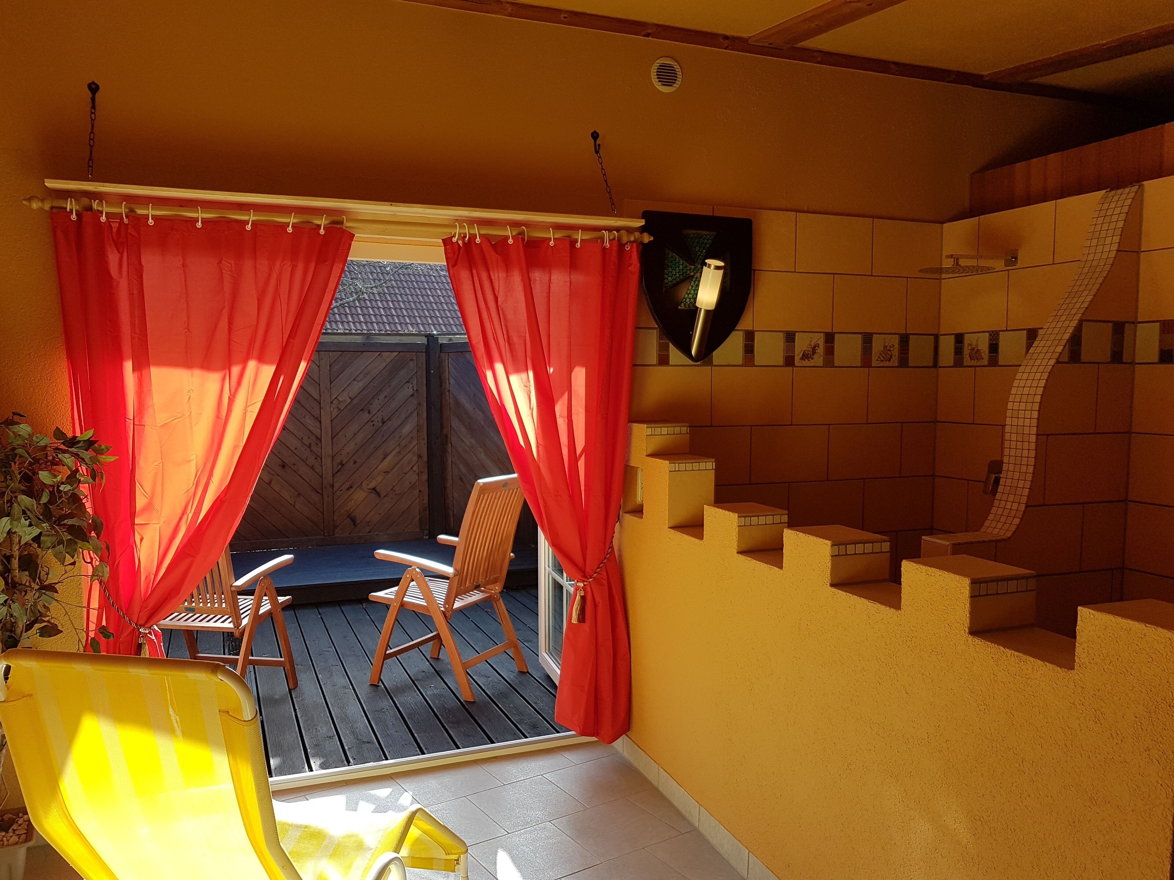A sunlit patio with a wooden deck, a yellow chair, and a red curtain.